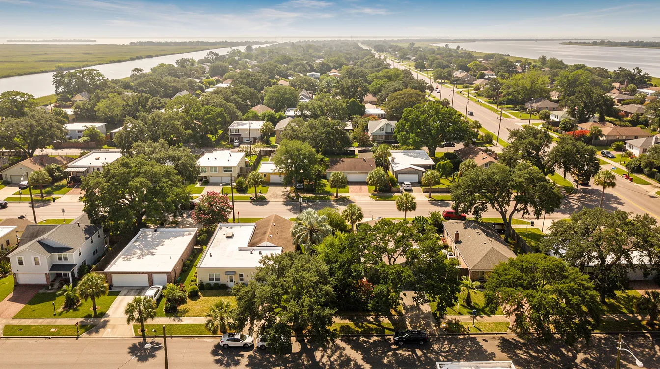An aerial view captures a Greater New Orleans suburban neighborhood on a hot summer day, showcasing flat rooftops and lush green trees. The scene emphasizes the importance of maintaining an efficient air conditioning system to combat the warm air and humidity typical of the region.